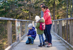 Eine glückliche Familie hält auf dem Baumwipfelpfad im Bayerischen Wald an, während ihr Kind ein Infoschild liest.