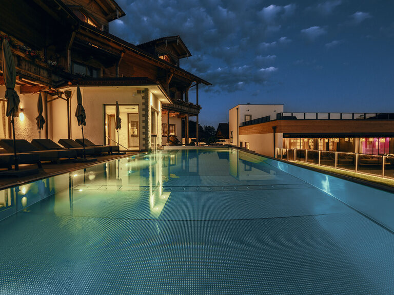 Blick auf den einladenden Pool des Tonihof mit mehreren Sonnenliegen und dem nächtlich beleuchteten Hotelgebäude im Hintergrund.